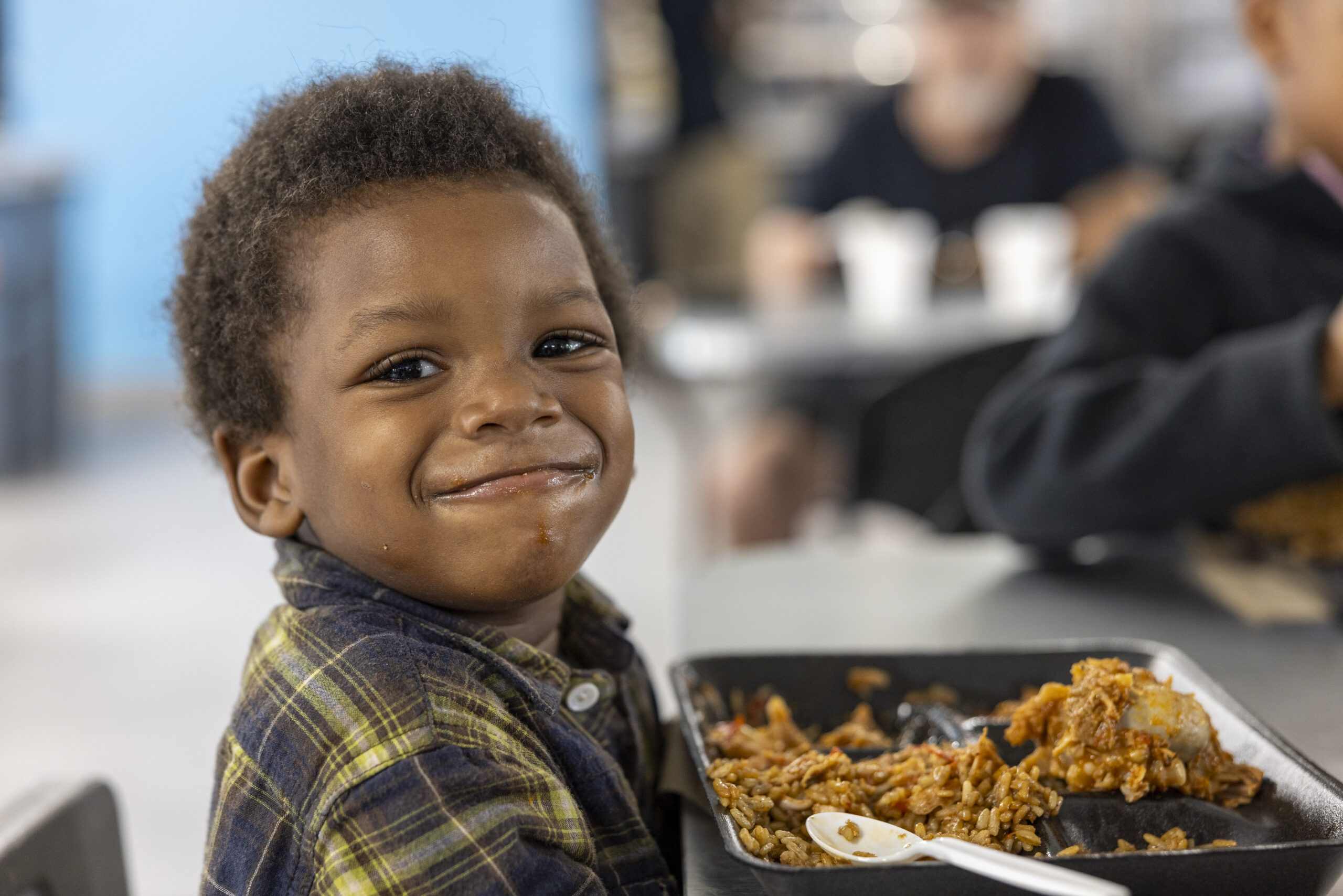 5 year old African american boy with a flannel shirt seated a table with a plate of food in front of him with.a mouth full of food and a grin